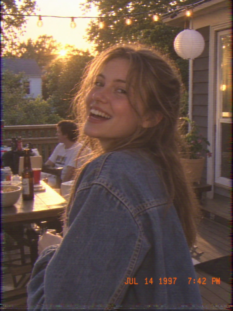 Hi8 camcorder style still of a laughing young woman in a denim jacket on a backyard porch at golden hour, with string lights and an orange date stamp reading JUL 14 1997